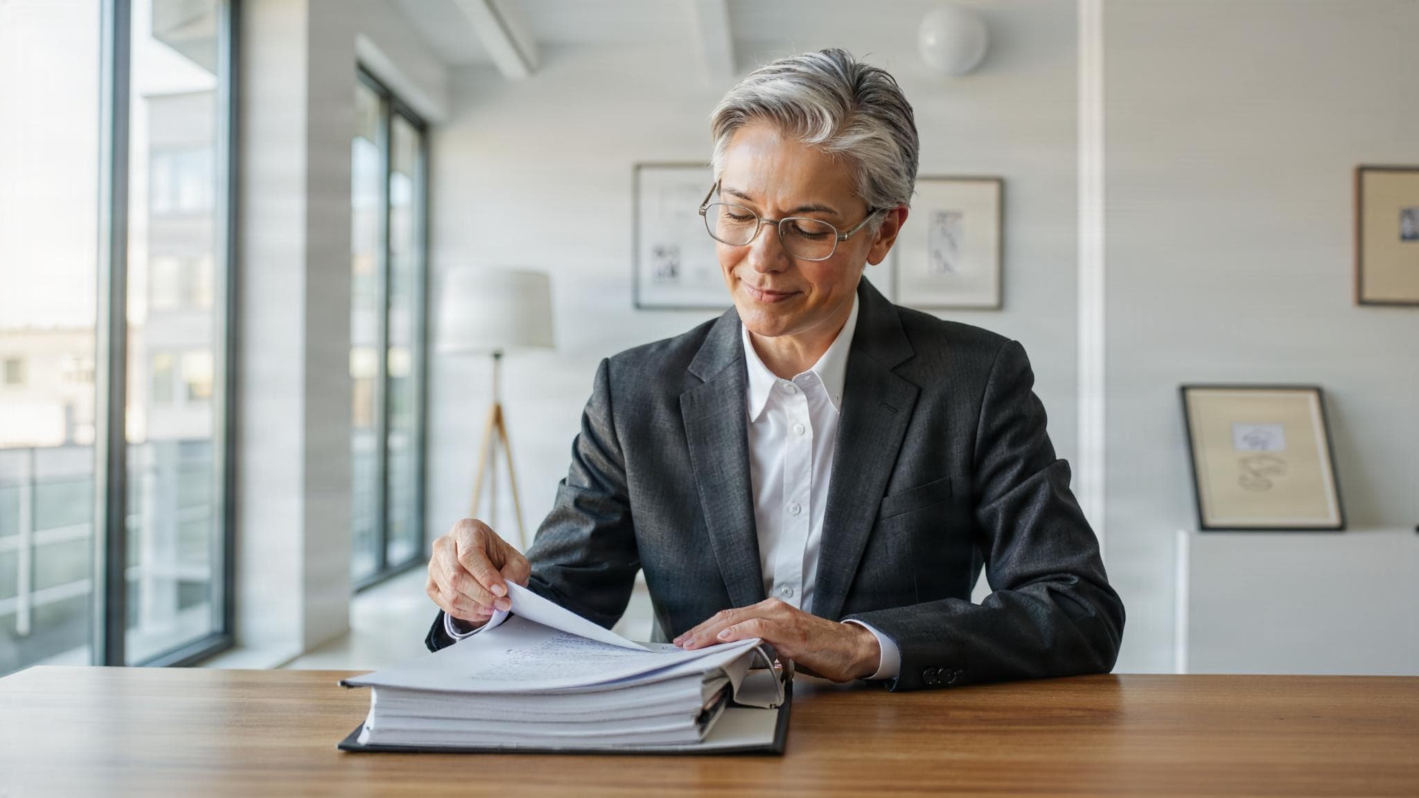 A grey-haired auditor leafs through a thick binder by the window, a certificate leaning against the wall.